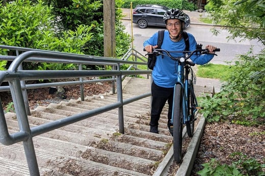 A photograph of Favyen Bastani, a man wearing a long-sleeved blue shirt, black pants, grey helmet and glasses, holding a bicycle while climbing stone stairs.